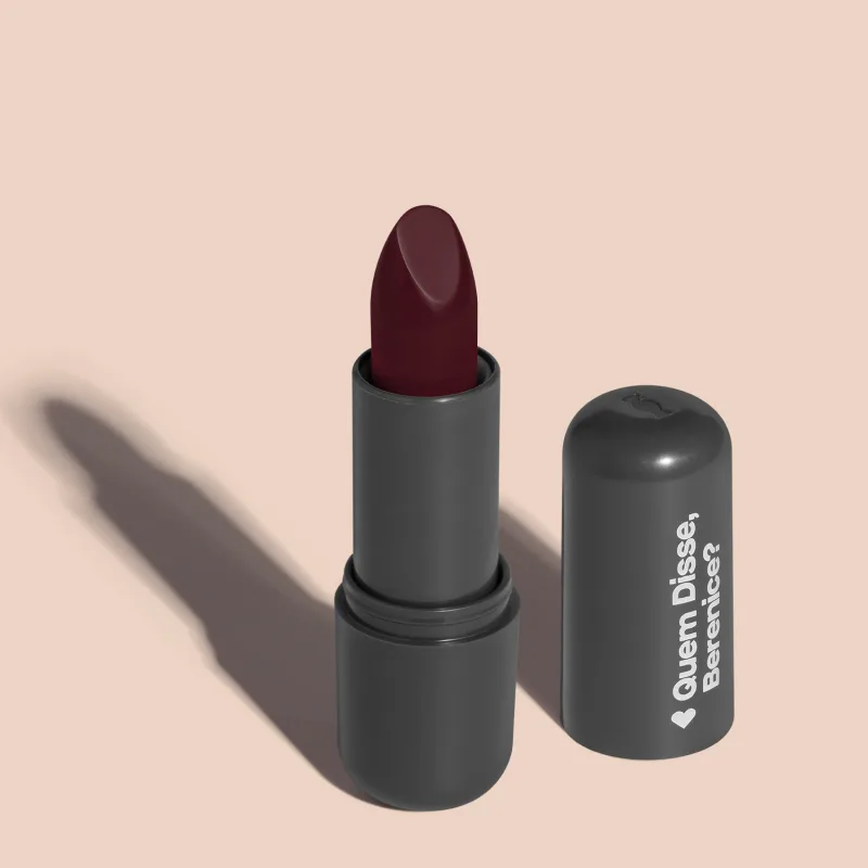 Batom de cor roxo fechado em embalagem preta com logotipo da marca Quem Disse, Berenice? em destaque. O batom está aberto sobre superfície rosa com iluminação que cria uma longa sombra projetada atrás da embalagem.