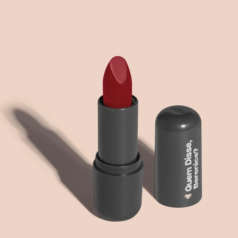 Batom de cor vermelho fechado em embalagem preta com logotipo da marca Quem Disse, Berenice? em destaque. O batom está aberto sobre superfície rosa com iluminação que cria uma longa sombra projetada atrás da embalagem.