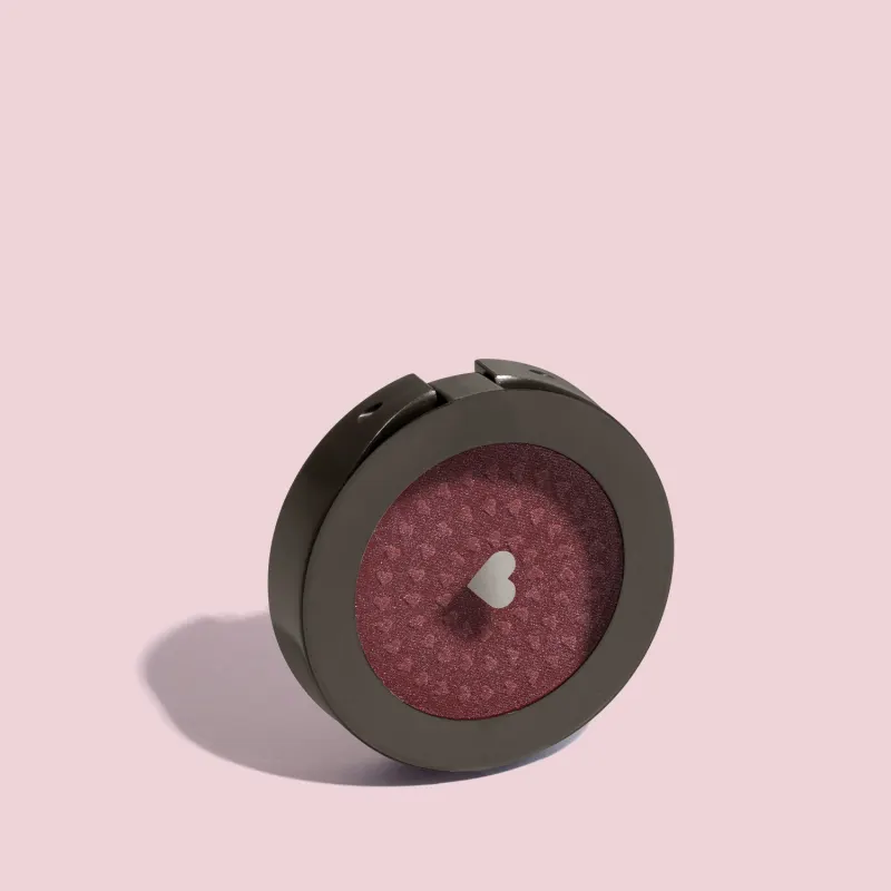 Estojo compacto redondo na cor preta com tampa transparente e coração ilustrado contendo 2g de blush em pó compacto Rubor Cobre Cobrelito da mara Quem Disse, Berenice? em fundo lilás.