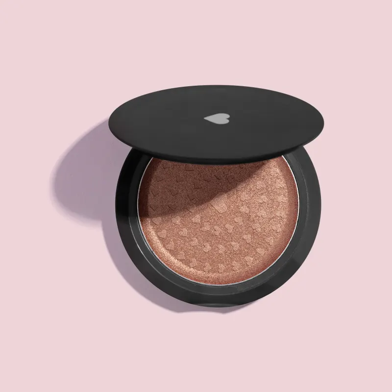Estojo compacto redondo na cor preta com tampa transparente e coração ilustrado contendo 5g de iluminador em pó compacto dourado Douradix Quem Disse, Berenice? em fundo lilás.