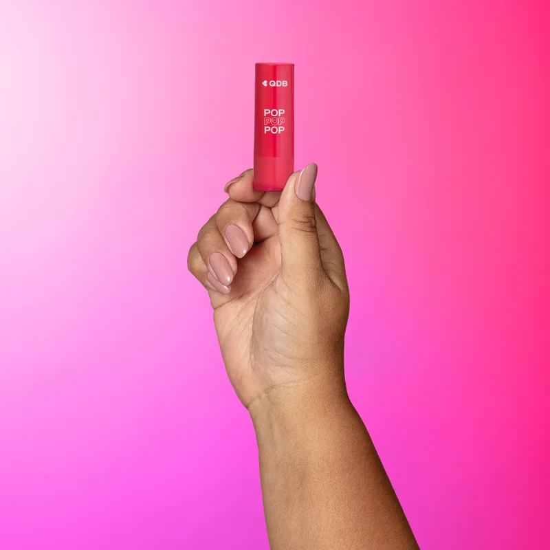 Imagem de uma mão segurando um batom da marca POP Quem Disse, Berenice? em embalagem rosa com letras brancas. O fundo tem um degradê de branco, roxo e rosa.