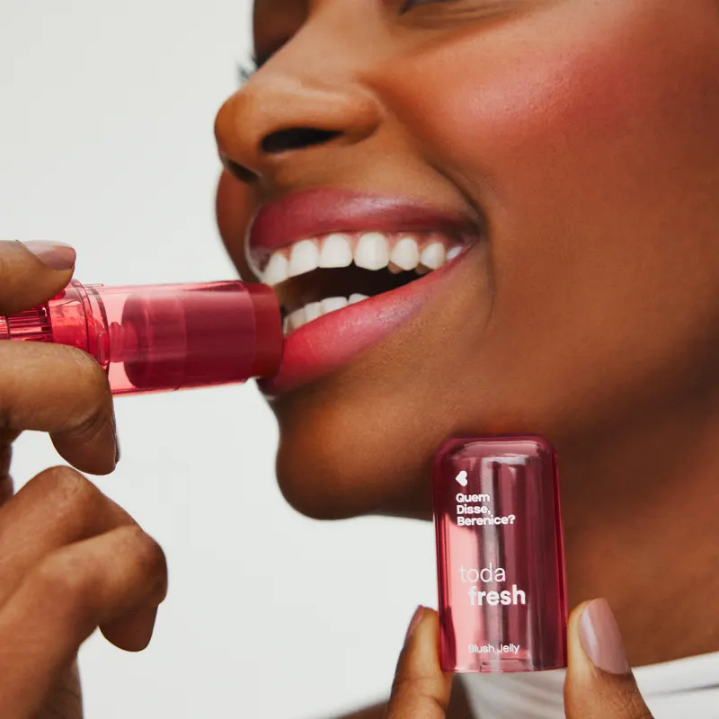 Uma mulher sorrindo passa blush jelly na cor pink da marca Toda Fresh Quem Disse, Berenice? no rosto com uma mão e a outra segura a tampa cor rosa do produto, em um fundo cinza claro.