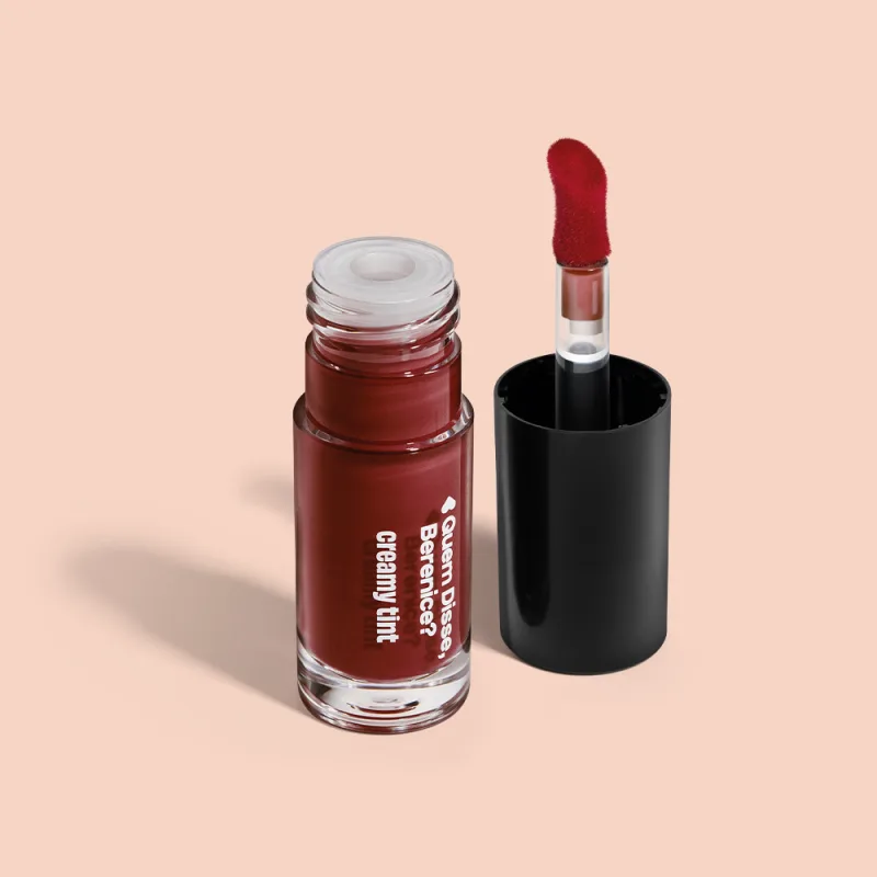 A imagem mostra um recipiente transparente de gloss labial da marca Quem Disse, Berenice? na cor vermelho cereja, com aplicador vermelho, haste transparente, tampa preta, sobre superfície clara e neutra.