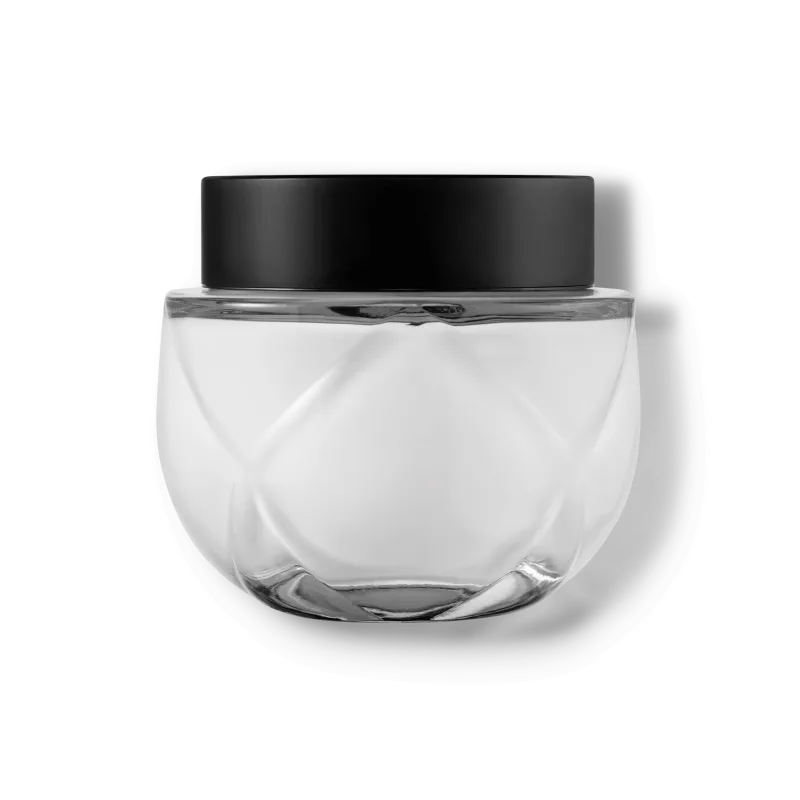 Frasco de vidro transparente arredondado com tampa preta. O recipiente tem uma forma esférica achatada. A tampa preta opaca contrasta com o vidro transparente do frasco que contém o creme na cor branca.