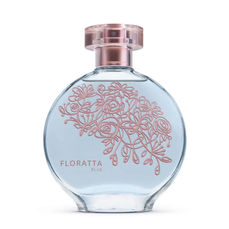 A imagem mostra um frasco de perfume da linha Floratta Blue. O frasco é de vidro transparente com um design arredondado. Há um padrão floral estilizado em tons de rosa desenhado sobre o fundo azul claro do frasco. A tampa é cor de rose gold metálico.