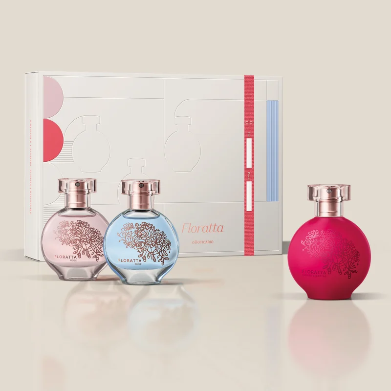 A imagem mostra uma linha de produtos de perfumaria da marca Floratta. Há três frascos de perfume em vidro transparente com detalhes gravados em rosa e azul claro, juntamente com uma caixa de papelão branca com elementos gráficos em rosa, vermelho e azul.