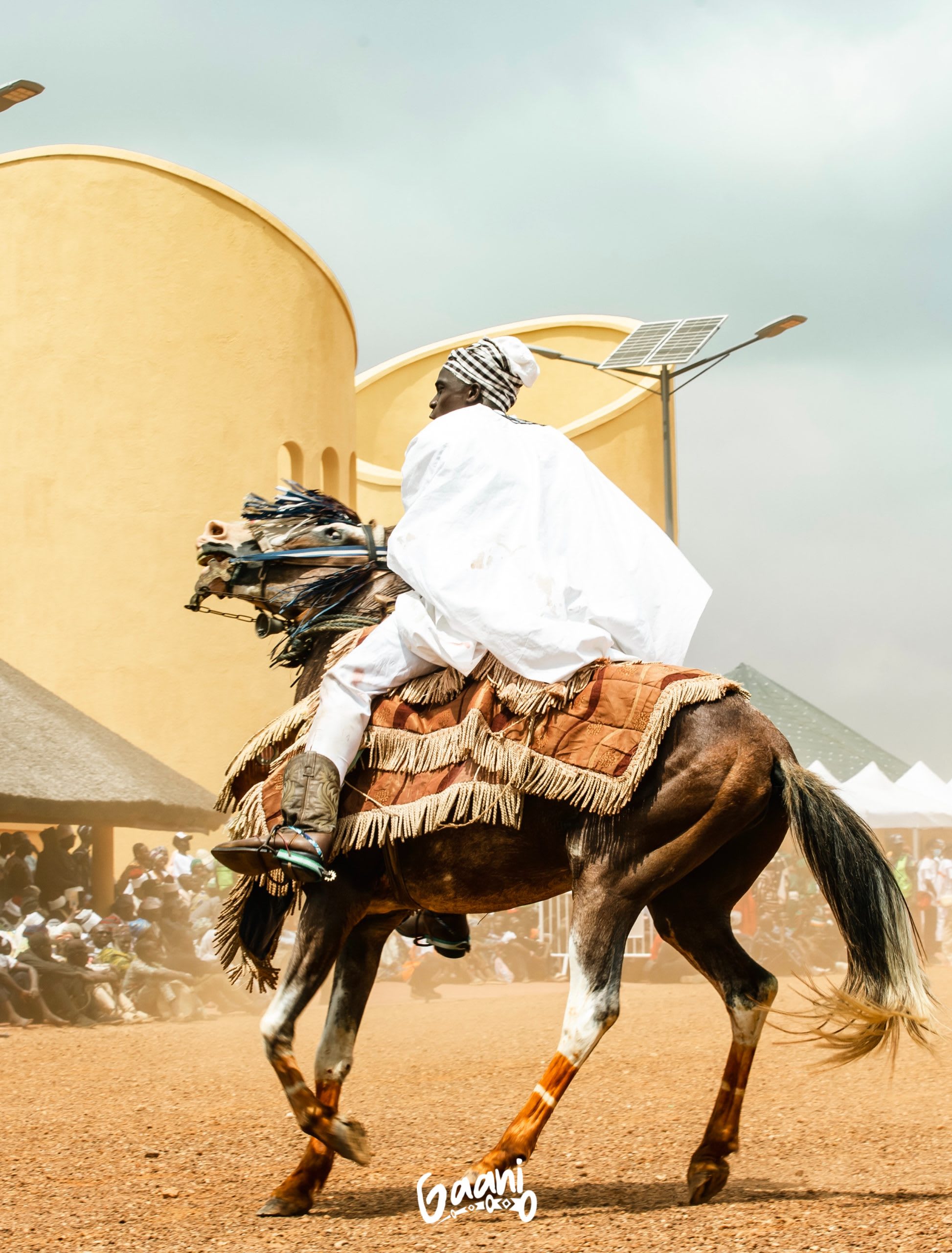 Cavalier en boubou blanc au galop devant un bâtiment cylindrique orné.