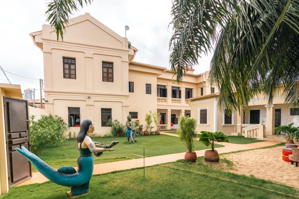 Grande maison crème à étage donnant sur un jardin, avec au premier plan une statue de sirène bleu-vert