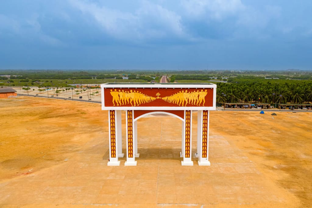 Vista aérea del monumento de la Puerta del No Retorno: un pórtico blanco adornado con un friso en relieve de siluetas doradas sobre un fondo marrón rojizo, en medio de un terreno de color ocre frente a una plantación de cocoteros