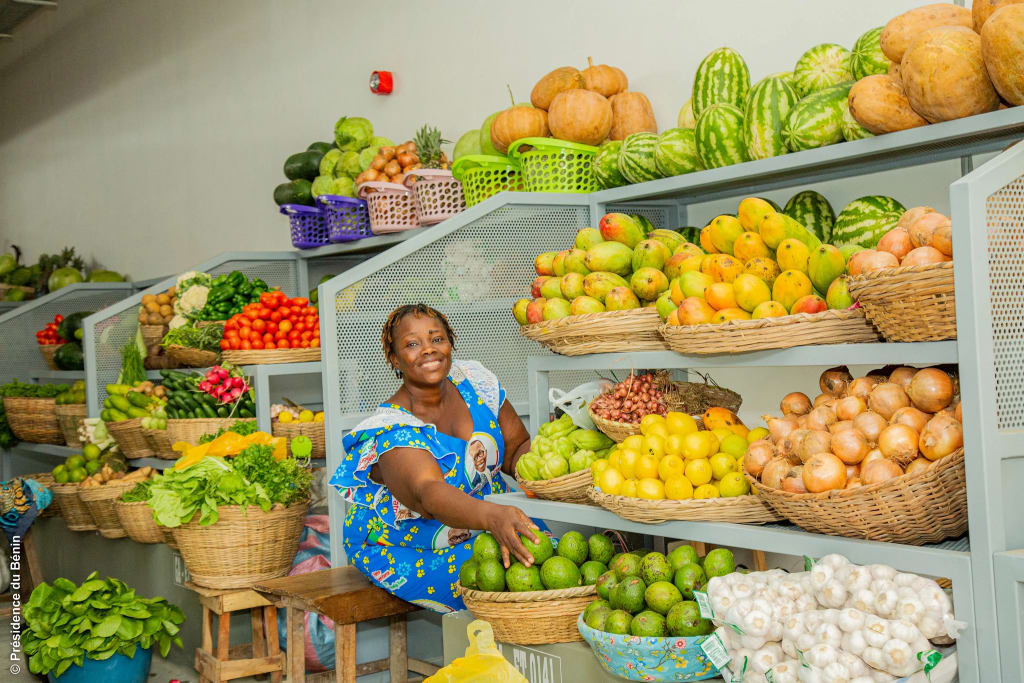 A smiling saleswoman, wearing a blue-and-yellow-patterned dress, sitting between shelves of fruit, vegetables, onions, and garlic, with one hand resting on some green fruit