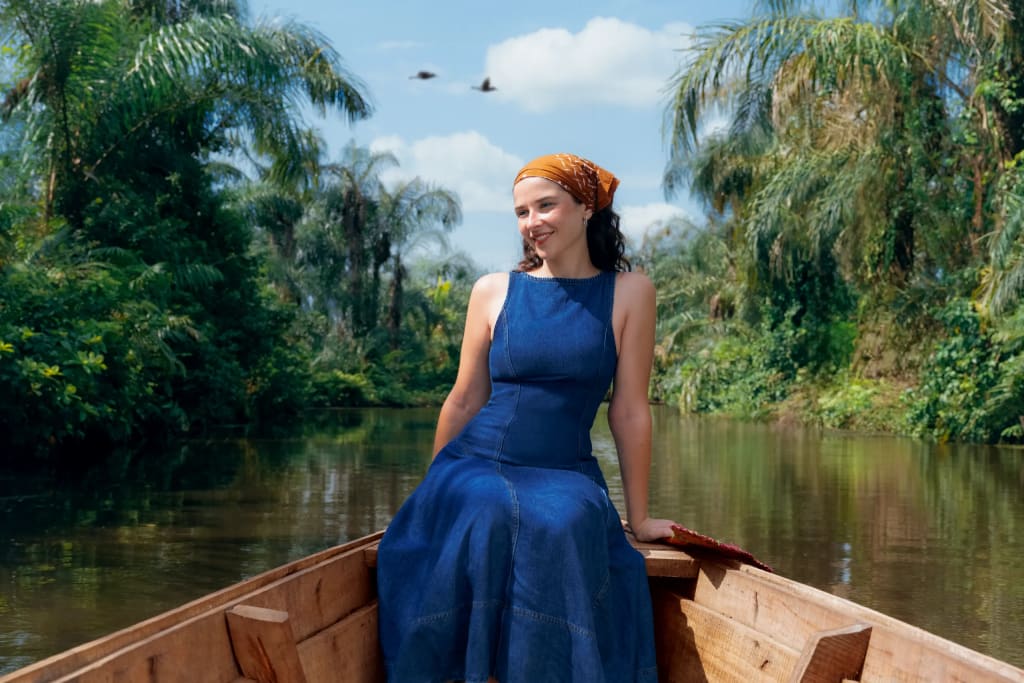 Une femme en robe bleue et foulard jaune assise à l’avant d’une pirogue, entre les raphias et les palmiers des deux rives