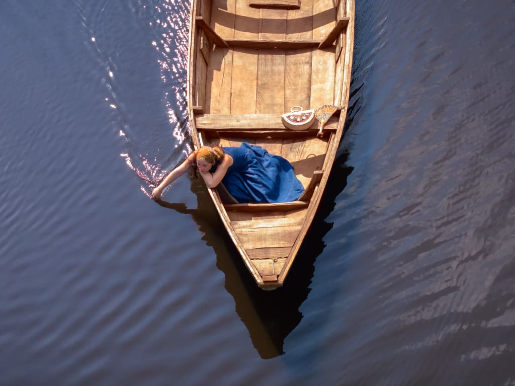 Vue du ciel d'une pirogue en bois glissant sur une eau sombre, une femme en robe bleue y laisse tremper sa main