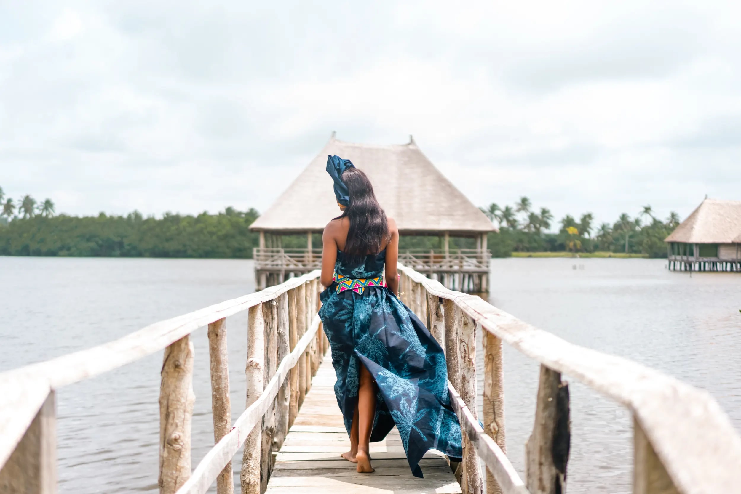 Une femme en robe bleu indigo marche de dos sur un ponton de bois au-dessus d’un lac, en direction de paillotes sur pilotis