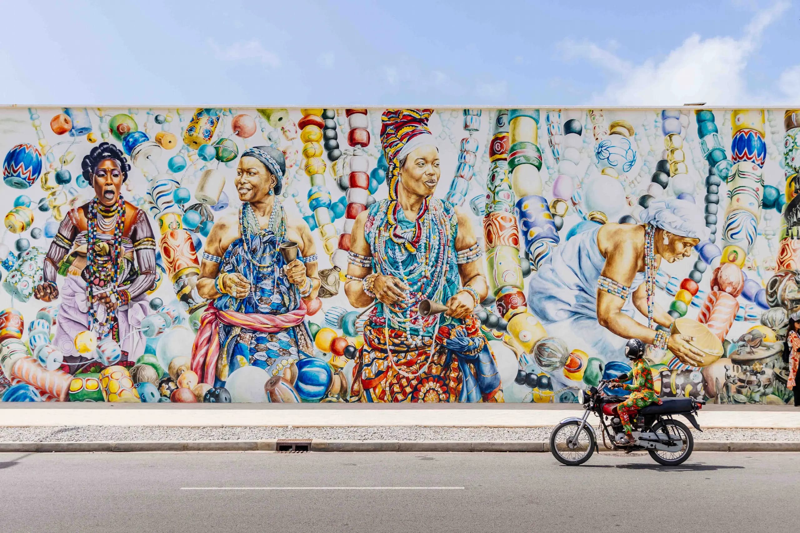 Immense fresque murale colorée représentant des femmes parées de perles et de pagnes, devant laquelle passe un motard