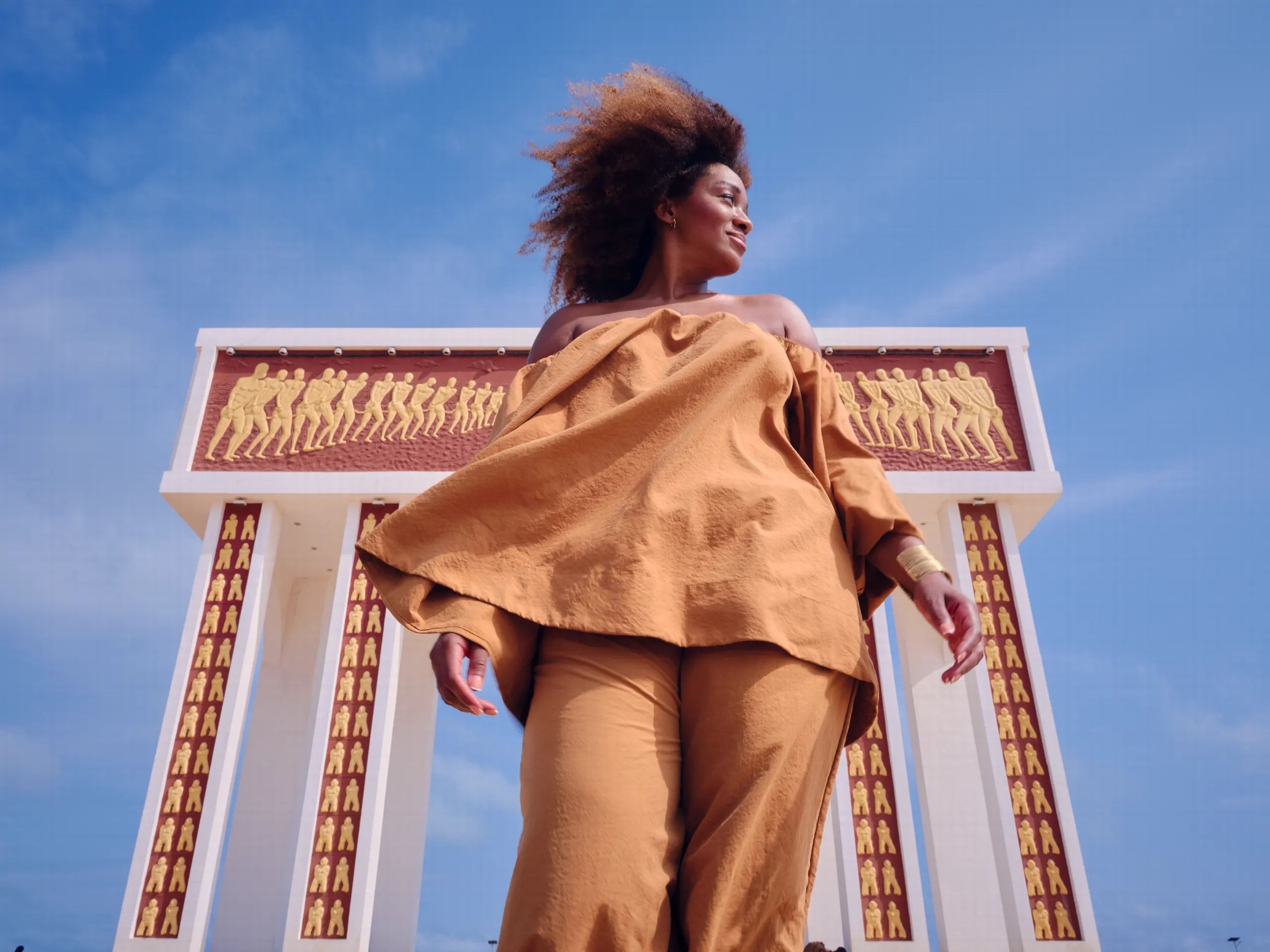 Una mujer vestida de beige, con el pelo al viento, vista desde abajo frente a un monumento blanco adornado con siluetas en relieve, bajo un cielo azul