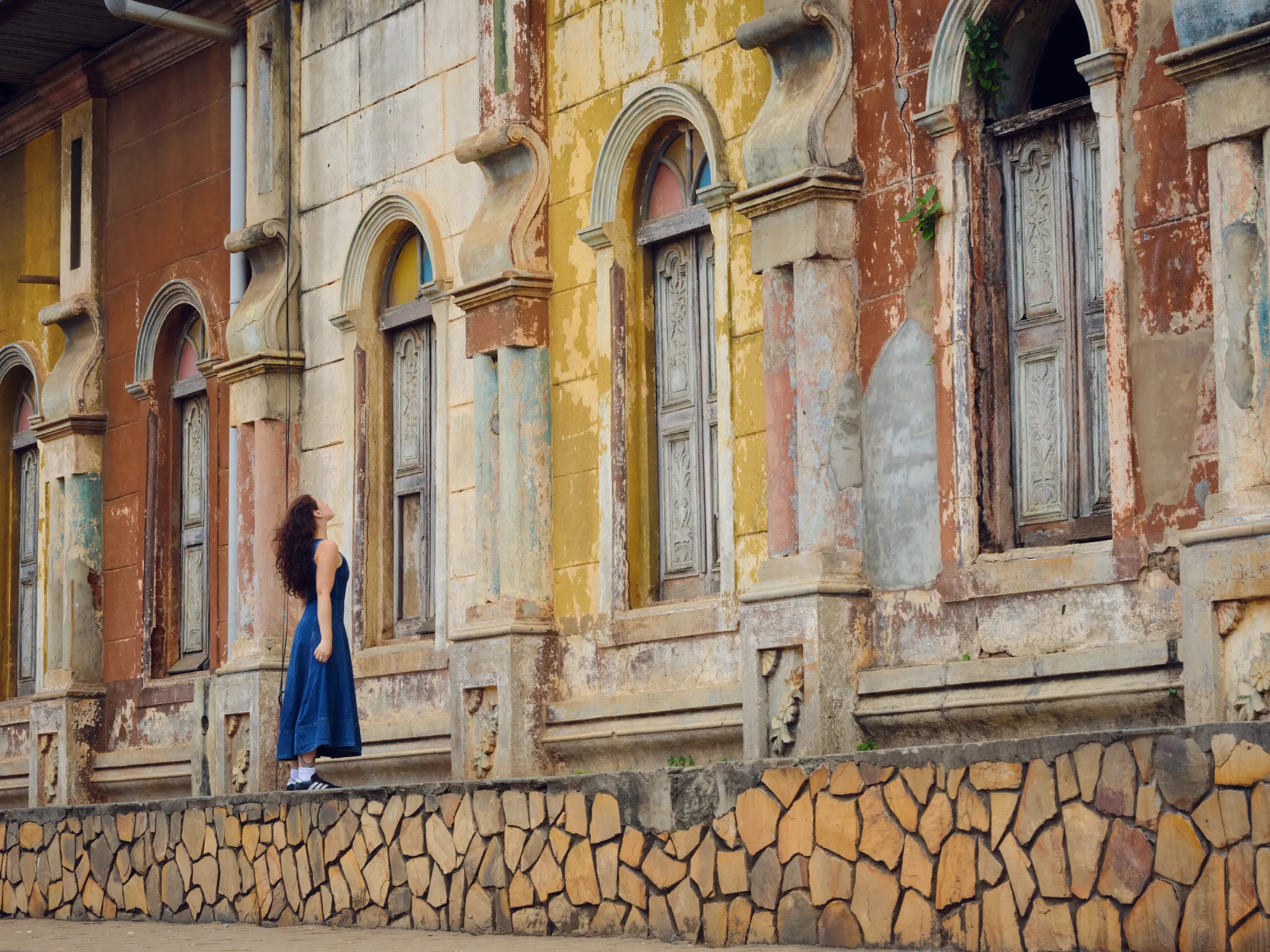 Une visiteuse en robe bleue lève les yeux vers d'anciens bâtiments aux façades patinées, jaunes et ocre, aux fenêtres cintrées et aux volets sculptés, à Porto-Novo