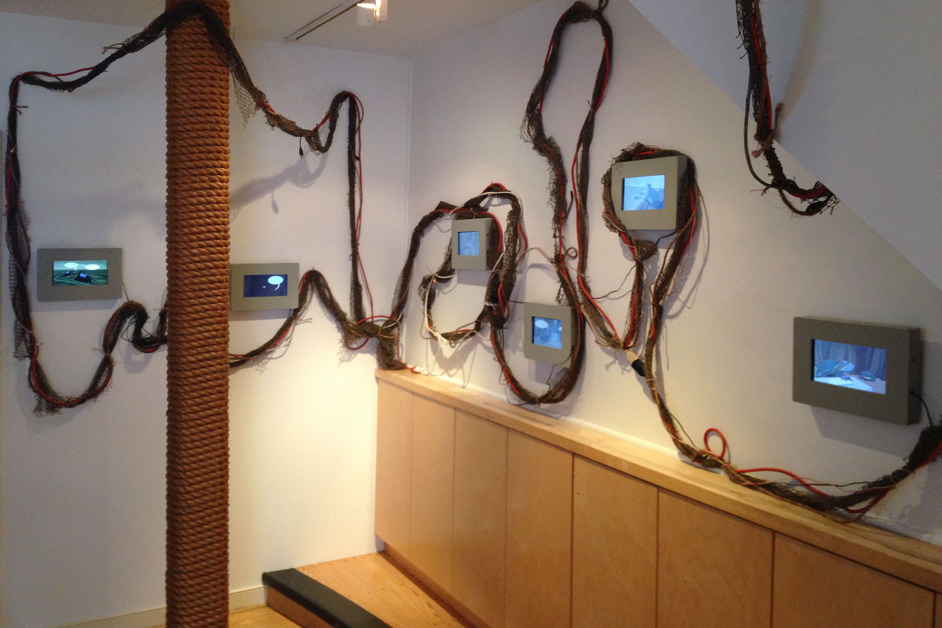 six small screens displayed on gallery walls with electrical cords and fishing net connecting them