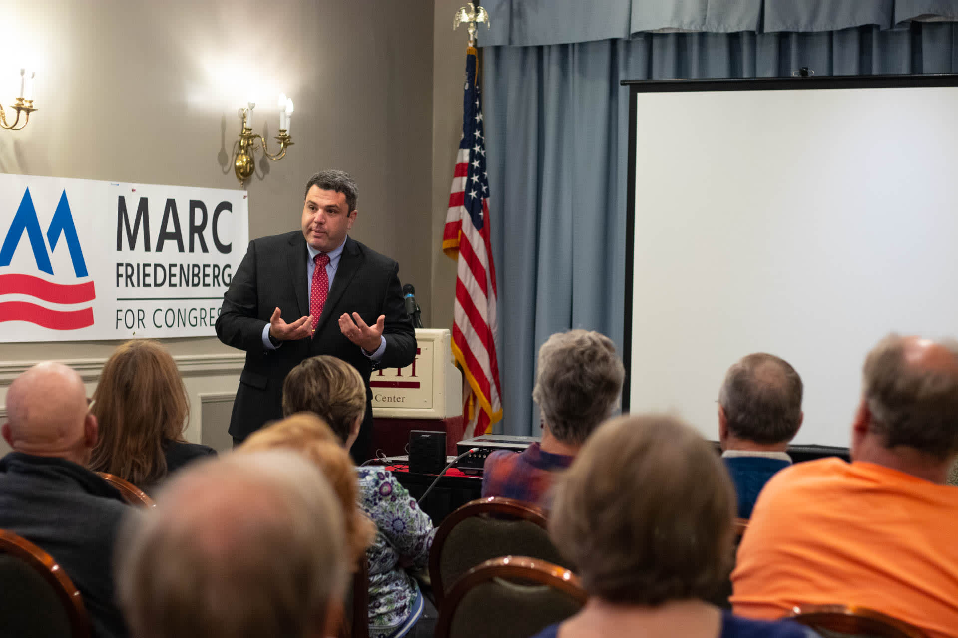 A man speaking to a crowd in a hotel lobby, with a large sign on the wall