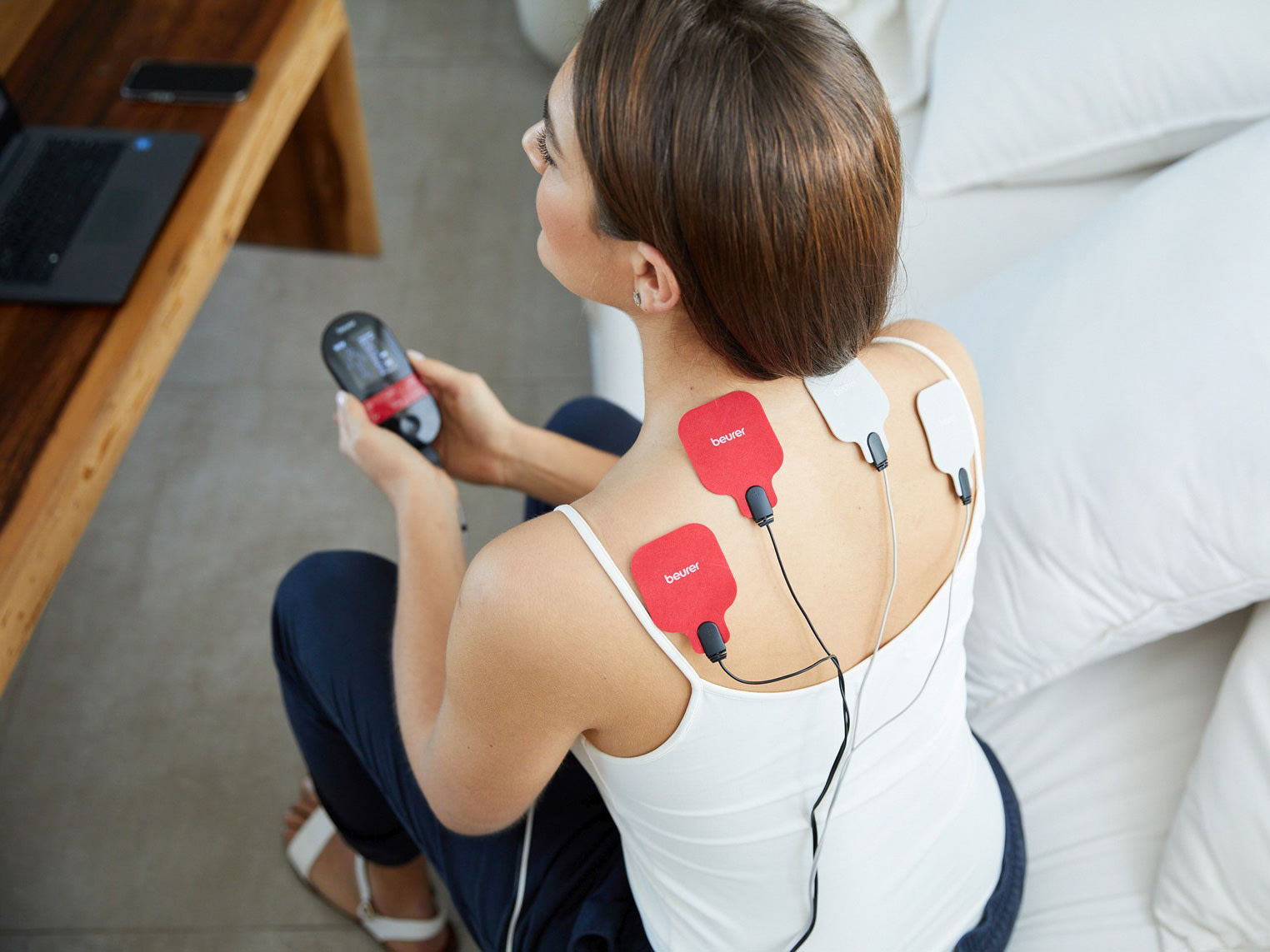 Frau sitzt auf dem Sofa und verwendet ein TENS-Gerät mit Elektroden im Schulter- und Nackenbereich.