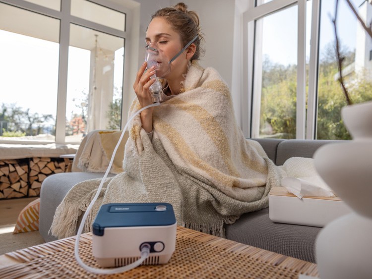 A woman is sitting on her sofa at home, using the Beurer IH 16 inhaler