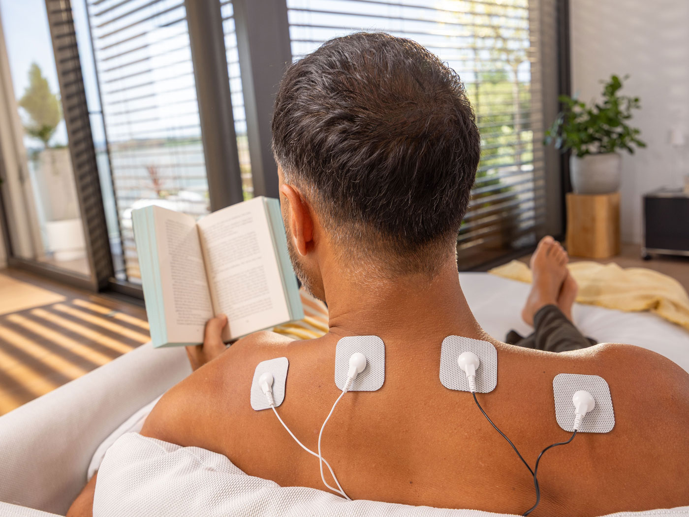 Mann liest auf dem Sofa, während Elektroden eines TENS-Geräts auf seinem oberen Rücken angebracht sind.