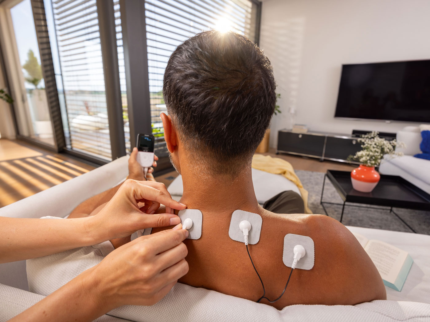 Hand platziert Elektroden für ein TENS-Gerät auf dem Nacken und oberen Rücken eines Mannes.