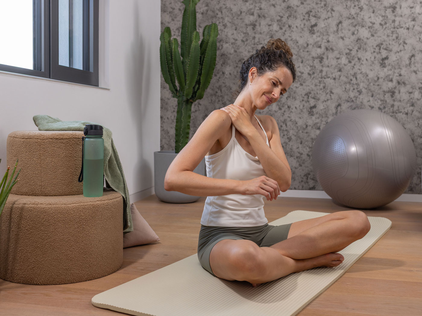 Frau sitzt auf einer Yogamatte und fasst sich wegen Nackenschmerzen an die Schulter.