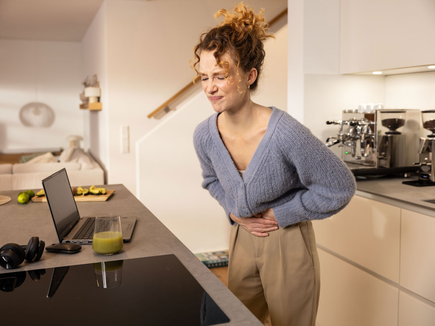 Frau hält sich mit beiden Händen den Unterbauch und steht mit Schmerzen in der Küche.