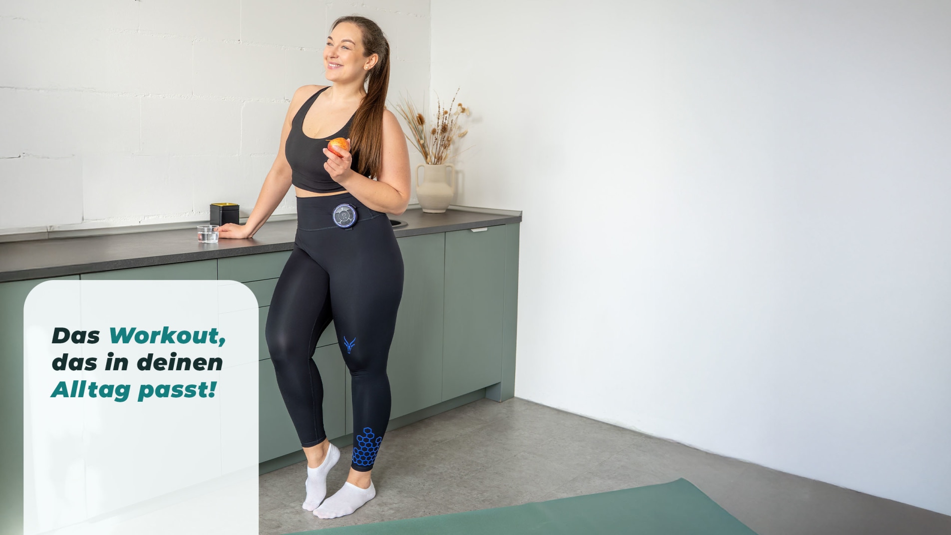 Eine junge Frau steht in der Küche mit einem Apfel in der Hand. Sie trägt die EMS High Waist Leggings von Antelope. Links neben ihr sieht man den Störer „Das Workout, das in deinen Alltag passt!“