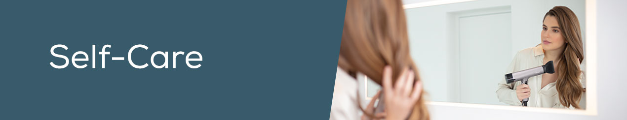 Banner for the “Self-Care” section: woman using the Beurer HC 100 hairdryer in front of a big, illuminated mirror