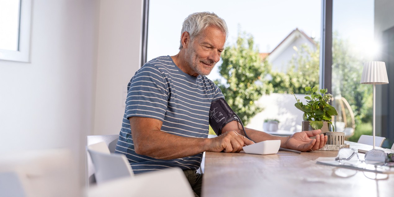 Un hombre mayor se toma la tensión arterial en el comedor con un tensiómetro Beurer.
