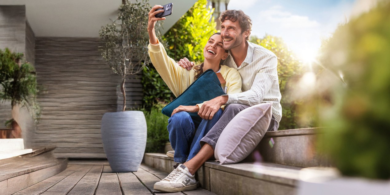 Junges Paar sitzt auf Terrasse im Garten und macht Sefie mit Beurer Heizkissen im Arm
