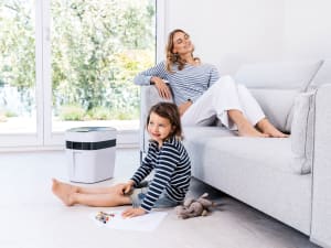 Mutter sitzt auf eine Sofa und Tochter daneben auf dem Boden und daneben steht das Beurer maremed Meeresklimagerät
