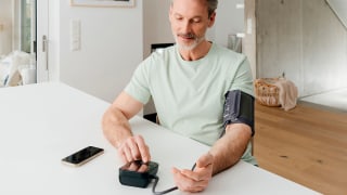 Elderly man is sitting at a table, measuring his blood pressure with the BM 48 upper-arm blood pressure monitor