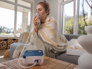 A woman is sitting on her sofa at home, using the Beurer IH 16 inhaler