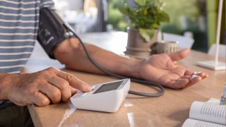 A person is measuring their blood pressure at the table using a Beurer upper-arm blood pressure monitor