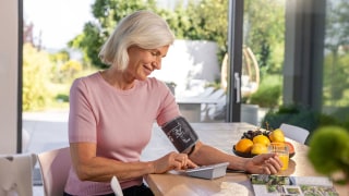 An elderly woman is sitting at a table, measuring her blood pressure with the BM 35 upper-arm blood pressure monitor