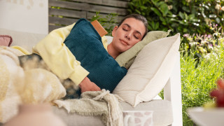 Una donna è distesa su una panchina in giardino con gli occhi chiusi e tiene tra le braccia il cuscino riscaldante a batteria HK 77