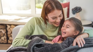 Mutter und Tochter sitzen lachend unter einer Wärmezudecke von Beurer auf dem Sofa