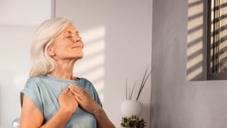 An elderly woman is standing at the window, her eyes closed, her hands resting relaxed on her chest