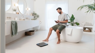 A man is sitting on the edge of the bath and controlling a Beurer diagnostic scale via his smartphone