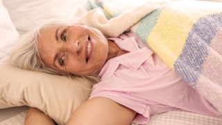 Ältere Frau liegt im Bett unter einer Decke und lächelt