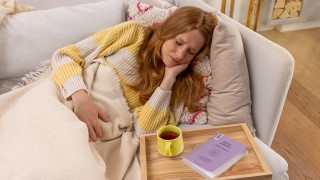 Frau liegt mit Unterleibsschmerzen auf der Couch und neben ihr steht eine Tasse Tee und liegt ein Buch