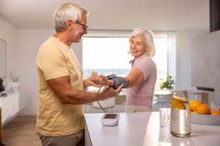 Frau steht mit Mann in der Küche und legt Oberarm-Blutdruckmanschette an