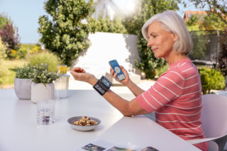 Frau misst ihren Blutdruck und kontrolliert gleichzeitig ihre Werte über die App "Beurer HealthManager Pro" an ihrem Smartphone