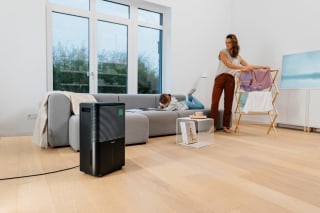 Ein Beurer LE 250 Luftentfeuchter steht im Vordergrund eines Wohnzimmers, während eine Frau Wäsche aufhängt und ein Kind auf dem Sofa liegt.