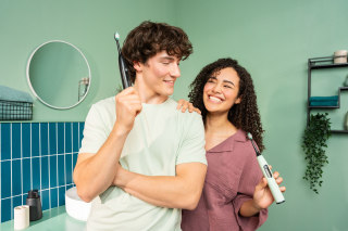Ein Mann hält eine schwarze elektrische Zahnbürste TB 50 im Badezimmer, während eine Frau im Hintergrund steht.