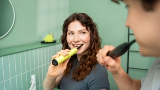 A woman brushing her teeth in the bathroom with the Beurer SC 50 electric toothbrush in the colour Splashy Lemon, next to her a man is also brushing his teeth.