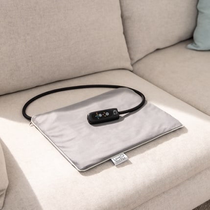 Graues Beurer-Wärmekissen aus der PetCare Serie liegt auf einem hellen Sofa.