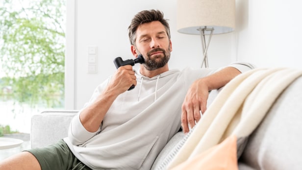 Mann massiert sich auf dem Sofa mit der Massagegun im Gesicht